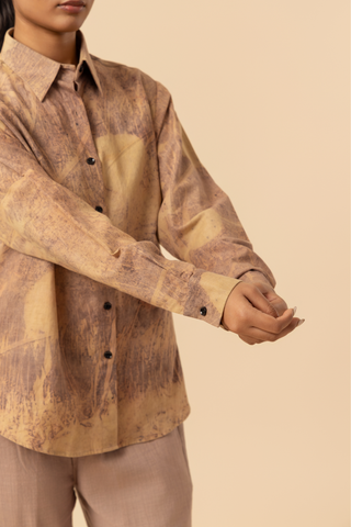 Closeup view of a model's hand stands against a light beige background, wearing a patterned co-ord set which has button-up shirt and wide-legged beige pants, accessorized with heels.
