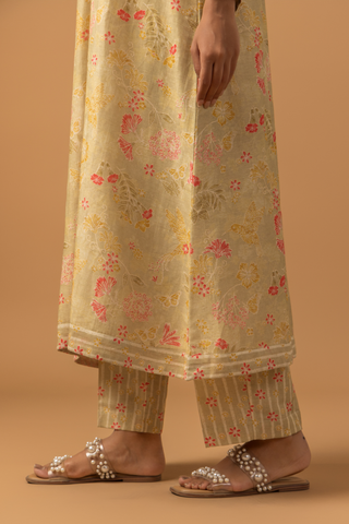 A woman in a floral-patterned, long beige linen co-ord set and striped pants, stands against a warm beige background.