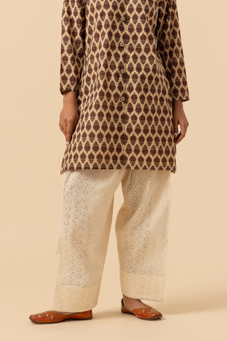 A person stands wearing a  traditional brown patterned chikan farshi salwar co-ord set with white embroidered pants and brown sandals against a neutral backdrop.