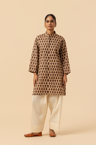 A person wearing a traditional brown patterned chikan farshi salwa  co-ord set with white embroidered pants and brown sandals, set against a neutral background.