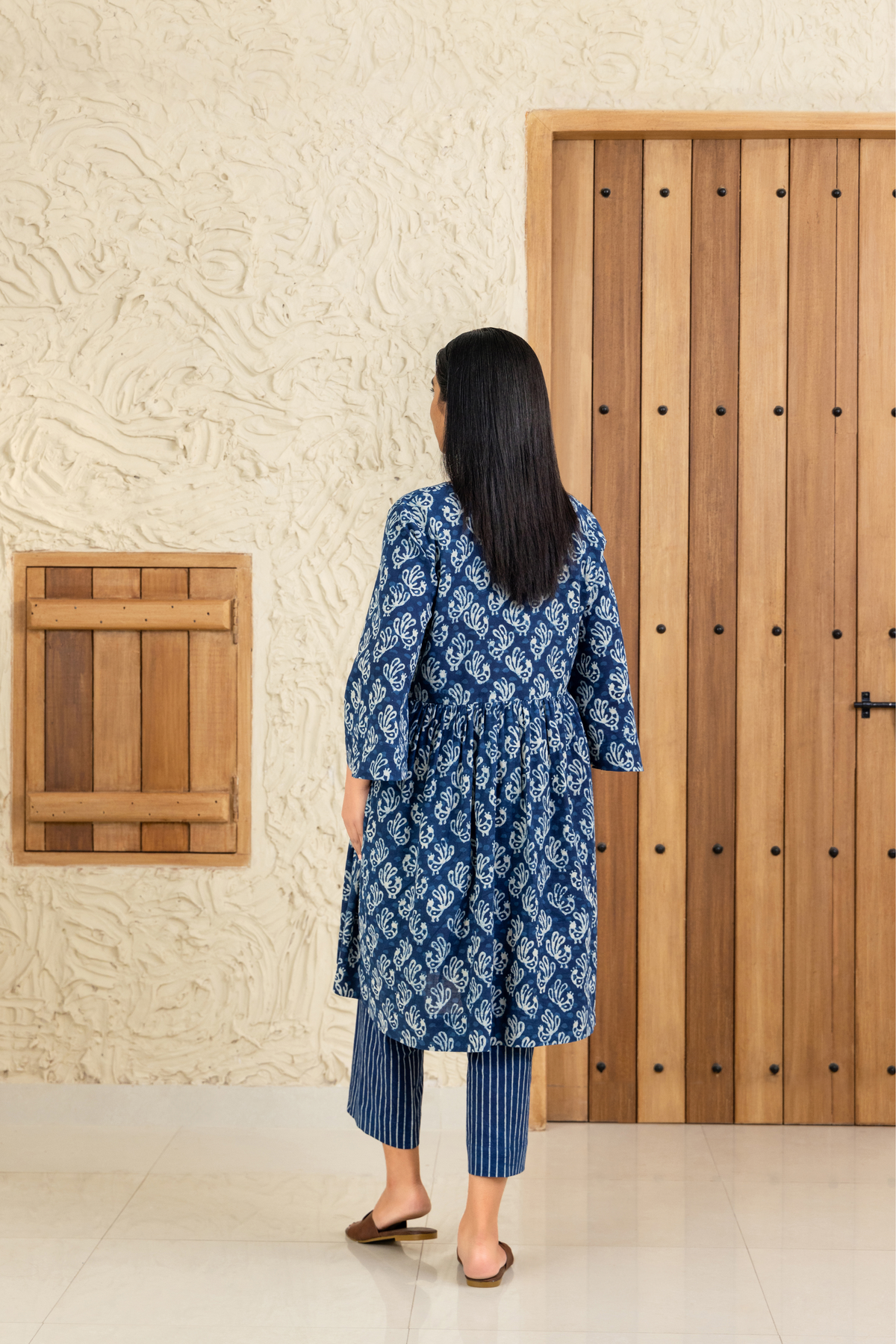 Back view of a woman wearing a blue and white printed co-ord set with a V-neck and tassel details, standing against a wooden and textured beige background.