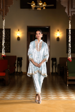 A woman wears a light blue ethnic velvet co-ord set outfit, standing gracefully in a spacious, elegantly decorated room with warm lighting and traditional furniture.