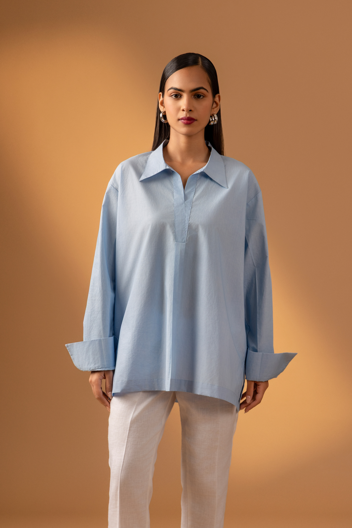 Closeup view of a women stands confidently wearing a light blue oversized shirt, white pants, and stylish sandals, against a warm beige background.