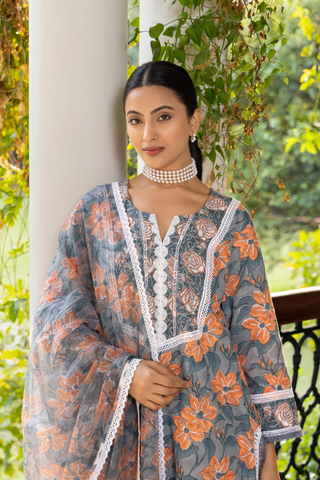 Closeup view of a woman stands elegantly in a floral, gray and orange cotton kurta set, complemented by a sheer scarf, amidst a lush green background.