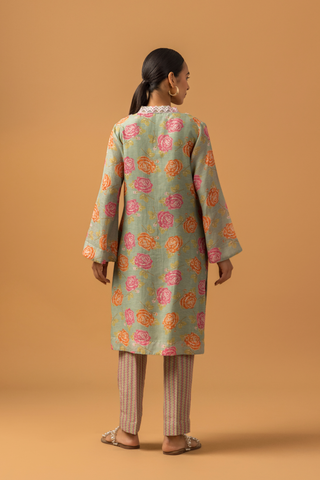 Back view of a model stands in a vibrant spring rose floral co-ord set with intricate neckline embroidery, paired with striped pants on a warm, neutral background.