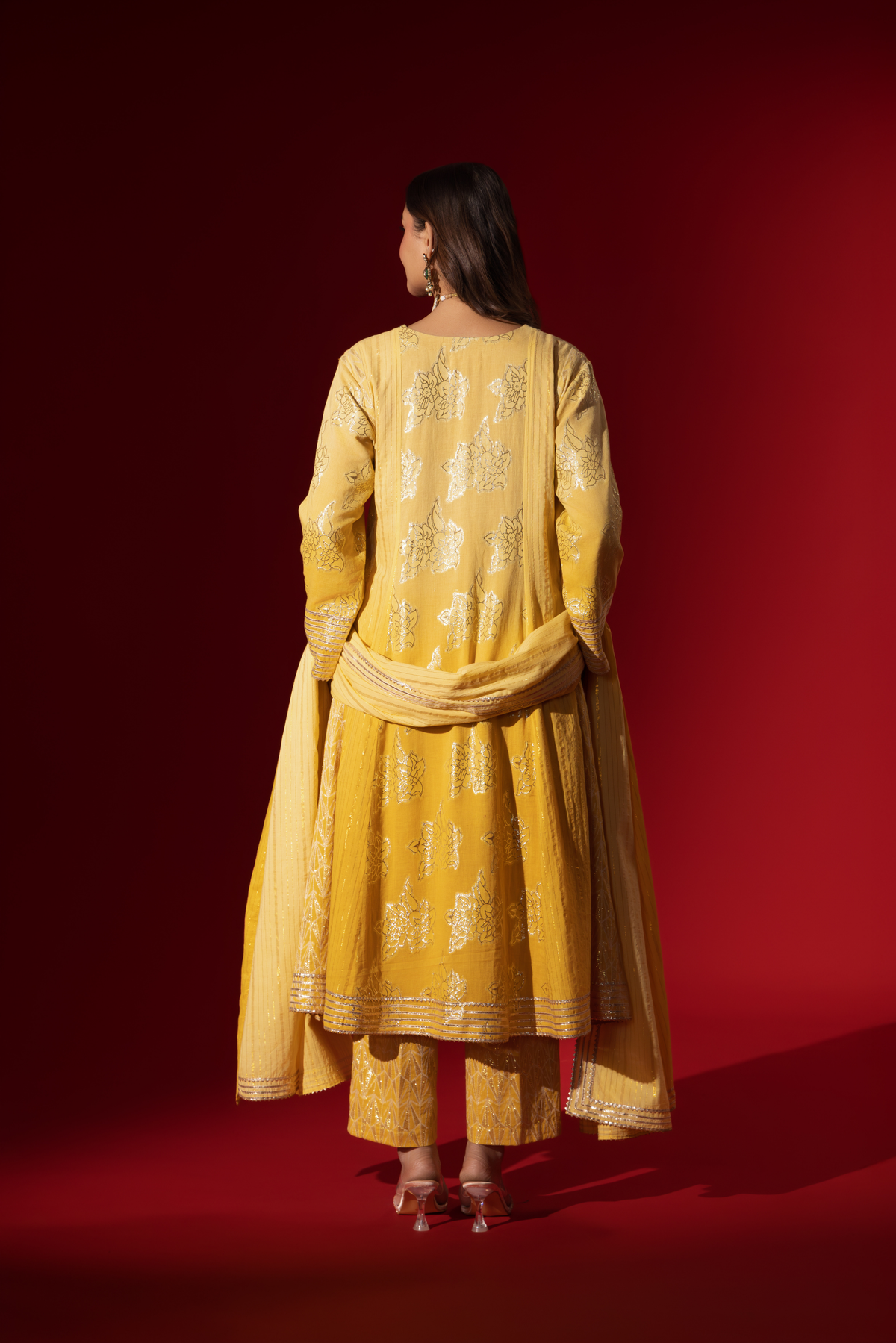 Back view of a woman showcases an elegant yellow traditional outfit with intricate designs, contrasting beautifully against a deep red background.