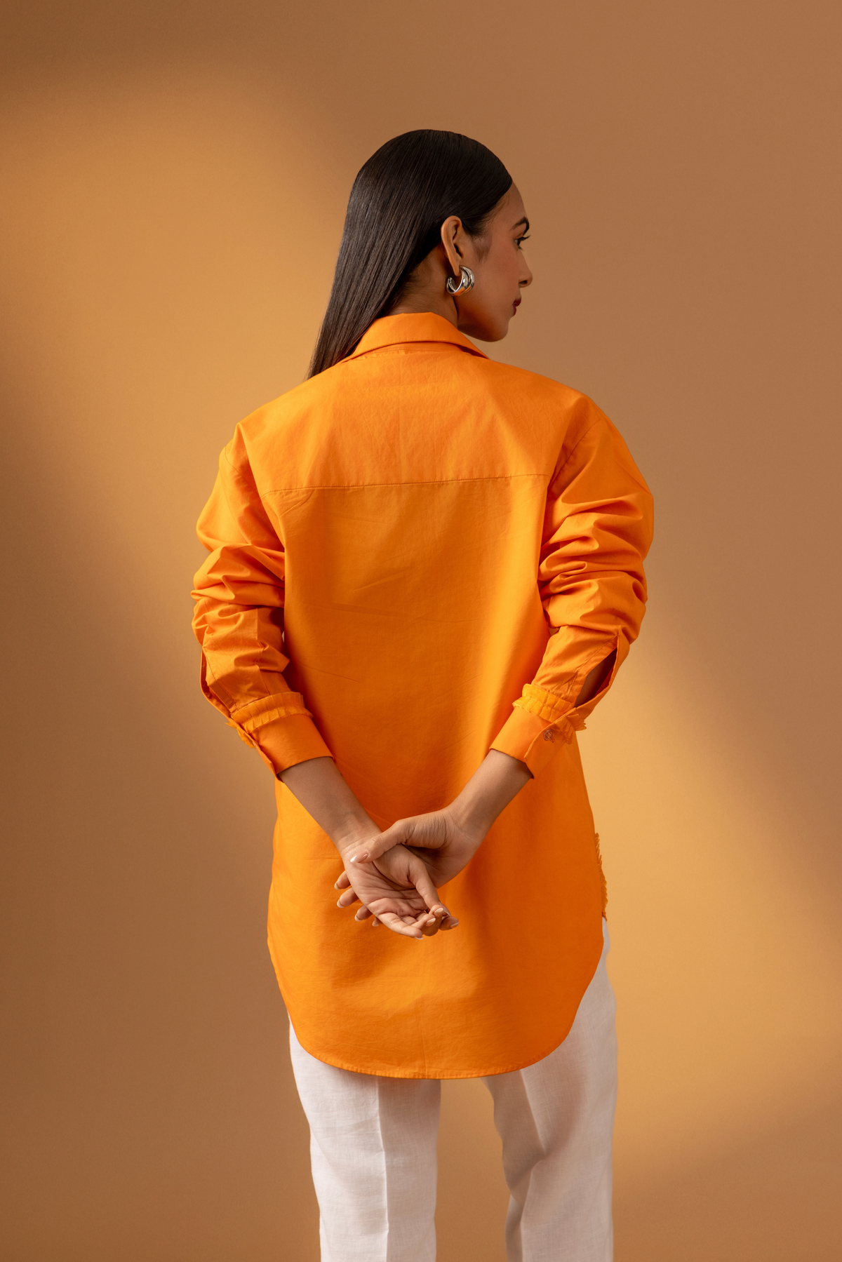 Back view of a woman stands confidently in an orange shirt paired with white trousers, and sandals, against a warm, neutral background.