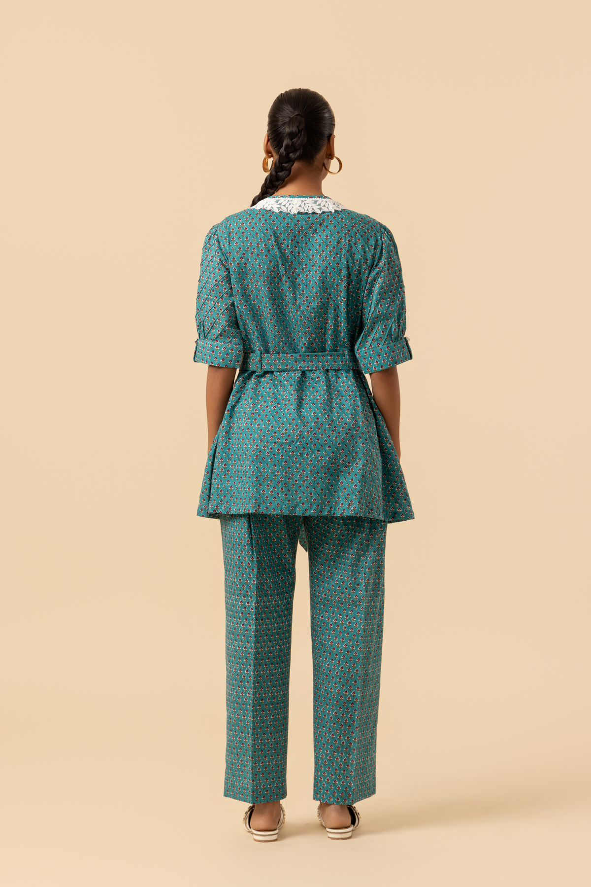 Back view of a person stands confidently, wearing a teal patterned outfit with a cinched waist and a collar, set against a neutral background.