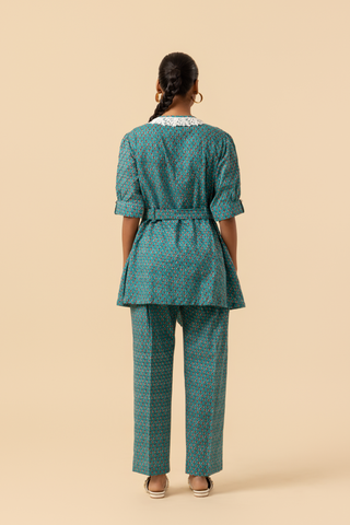 Back view of a person stands confidently, wearing a teal patterned outfit with a cinched waist and a collar, set against a neutral background.