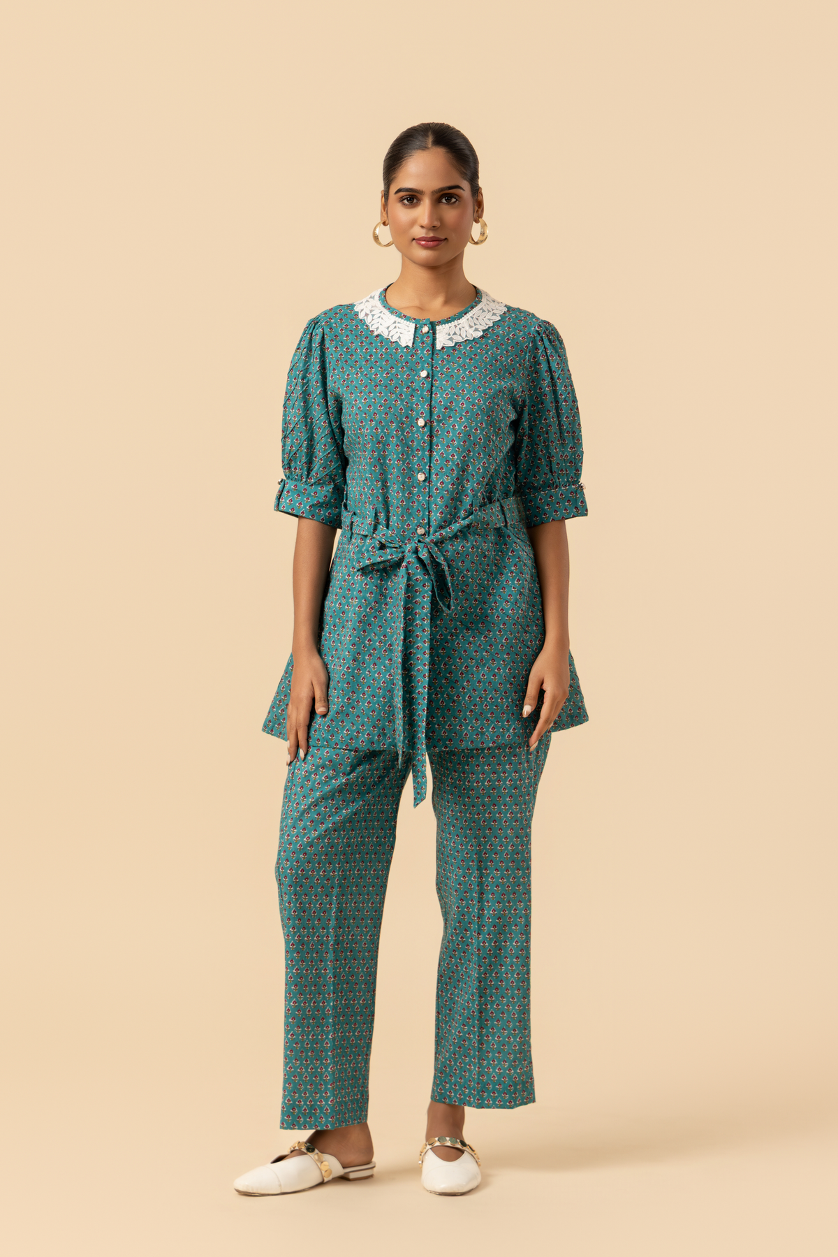 A person stands confidently, wearing a teal patterned outfit with a cinched waist and a collar, set against a neutral background.