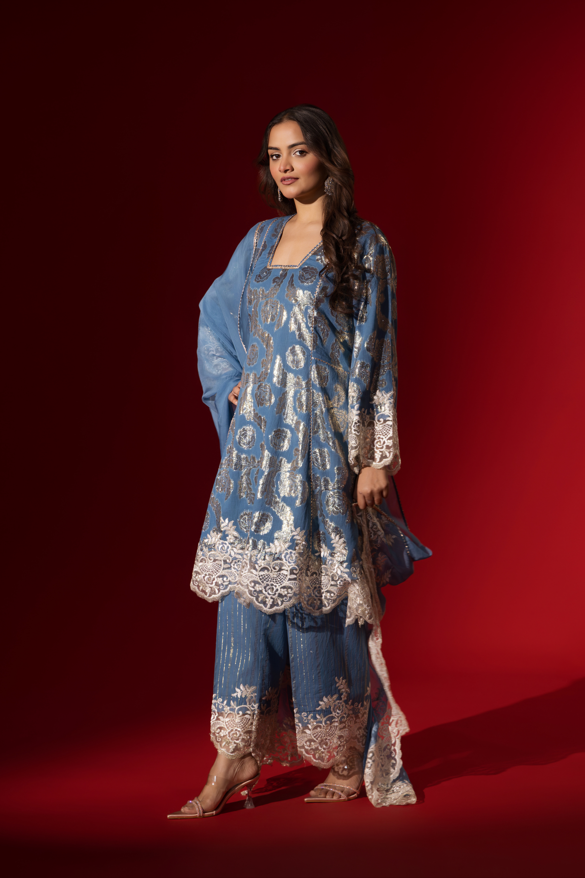 Side view of a woman poses in a blue ethnic  shimmer kurta set outfit with intricate white embroidery against a vibrant red backdrop.