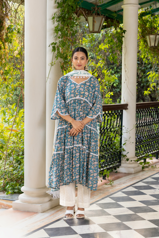 A woman stands gracefully in a patterned blue kurta set and white pants, in a lush green setting with ornate pillars and checkered flooring.