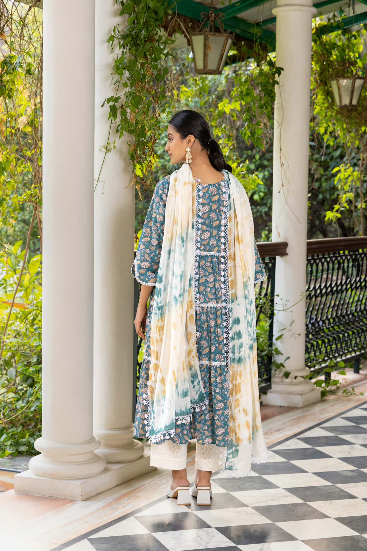 Back view of a woman stands gracefully in a patterned blue kurta set and white pants, in a lush green setting with ornate pillars and checkered flooring.