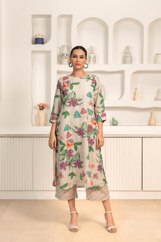 A woman is standing indoors wearing a light-colored floral-printed kurta set paired with patterned pants. She has her hair tied back and accessorized with hoop earrings. She is posing against a minimalist white background featuring built-in shelves decorated with vases and small decorative items.