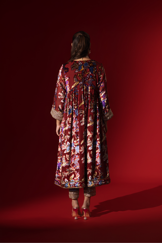 Back view of a woman in traditional attire features a richly embellished maroon and floral dress, paired with a blue shawl, against a deep red background.