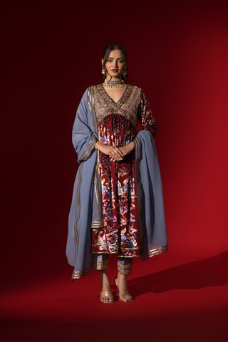 A woman in traditional attire features a richly embellished maroon and floral dress, paired with a blue shawl, against a deep red background.