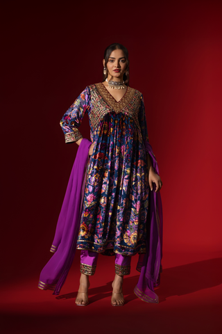 A woman stands confidently in a vibrant, embroidered purple printed velvet anarkali kurta set, paired with a matching dupatta against a rich red backdrop.