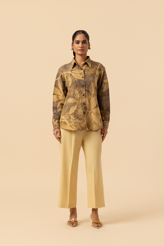 A model wearing a patterned brown and yellow shirt with beige wide-leg trousers and sandals, standing against a soft beige background.