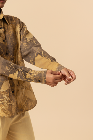 Closeup view of a woman's hand wearing a patterned brown and yellow shirt with beige wide-leg trousers and sandals, standing against a soft beige background.