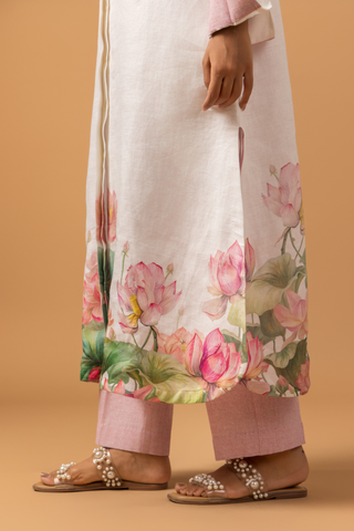 A woman in a floral-printed kurta with pink lotus designs and matching pants, paired with embellished sandals on a warm background.