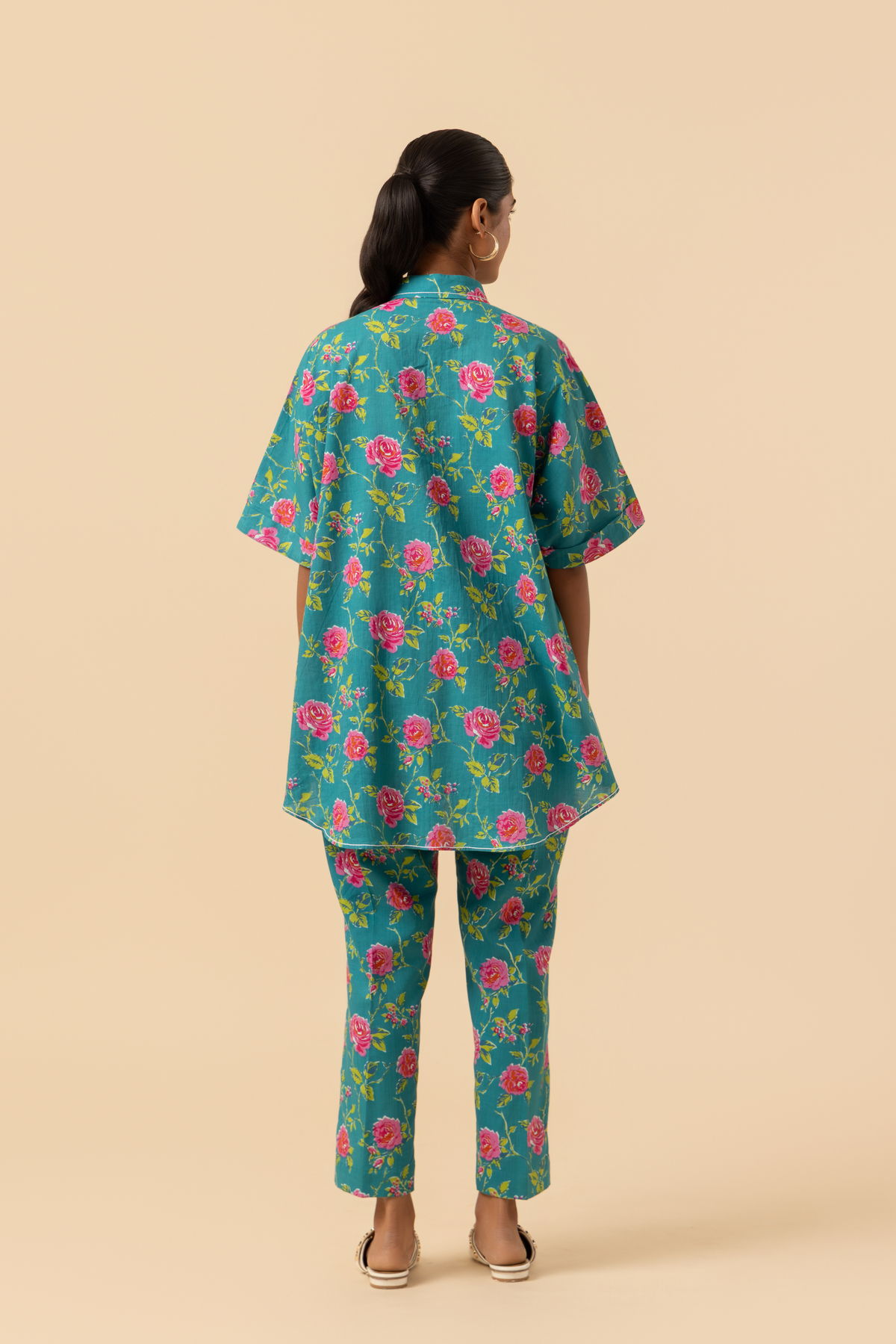 Back view of a person models a vibrant floral outfit, featuring a teal shirt and matching pants adorned with pink roses, set against a neutral background.