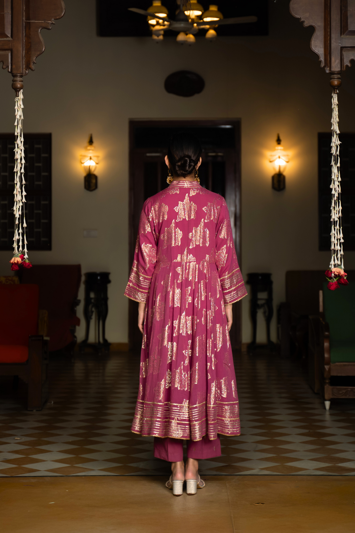 Back view of a  woman in a dark pink traditional anarkali kurta with intricate gold patterns, standing in a beautifully decorated interior.