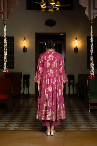 Back view of a  woman in a dark pink traditional anarkali kurta with intricate gold patterns, standing in a beautifully decorated interior.