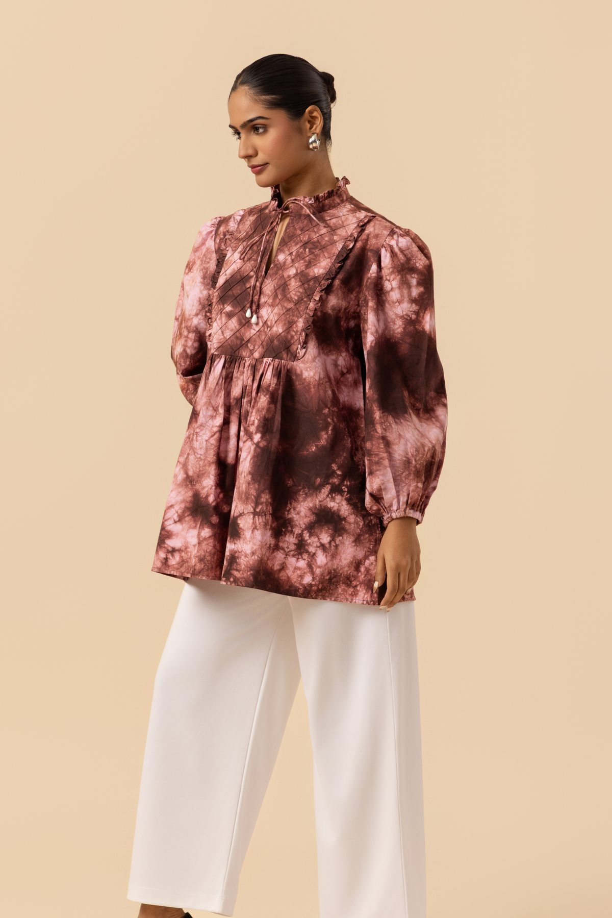 Side view of a model wears a vibrant brown tie-dye shirt dress with puffed sleeves, paired with white wide-leg trousers and stylish sandals.