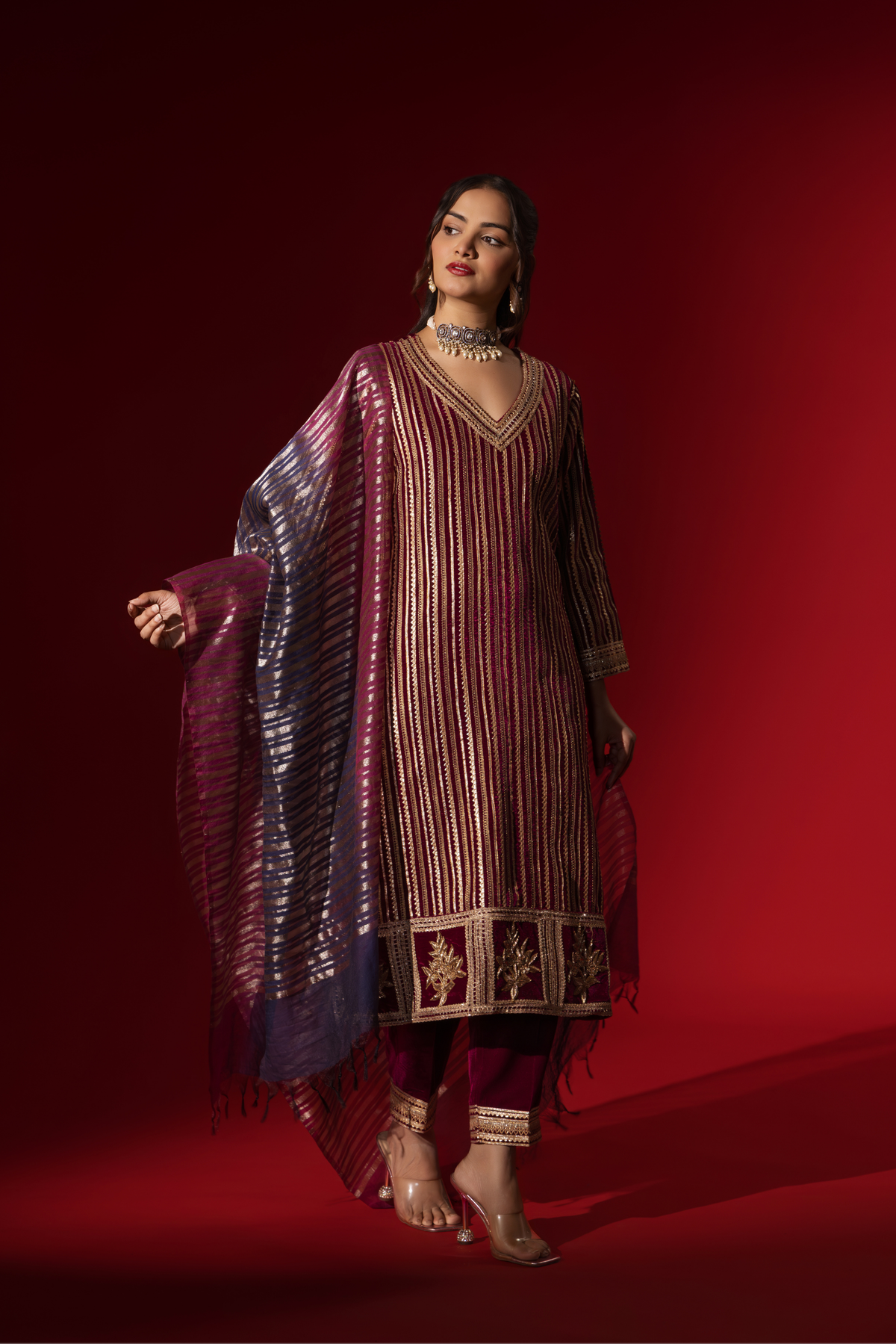 Side tilt view of a woman showcases an ethnic wine velvet gota kurta set with golden stripes, paired with a shimmering shawl, against a rich red backdrop.