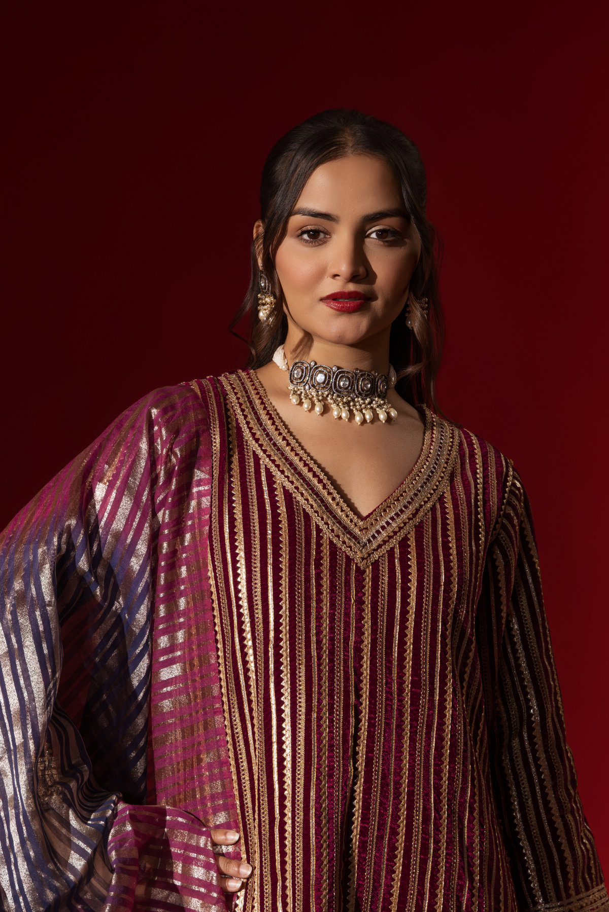 Closeup view of a woman showcases an ethnic wine velvet gota kurta set with golden stripes, paired with a shimmering shawl, against a rich red backdrop.