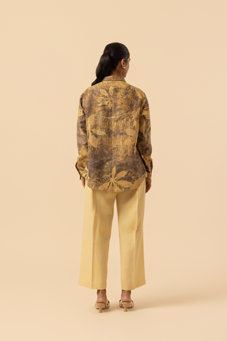 Back view of a model wearing a patterned brown and yellow shirt with beige wide-leg trousers and sandals, standing against a soft beige background.
