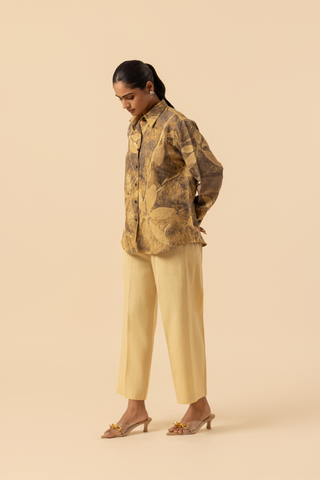 Side view of a model wearing a patterned brown and yellow shirt with beige wide-leg trousers and sandals, standing against a soft beige background.