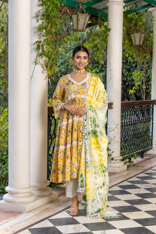 A woman stands gracefully in a vibrant yellow floral cotton kurta set, paired with a light green and white dupatta against a lush, green backdrop.