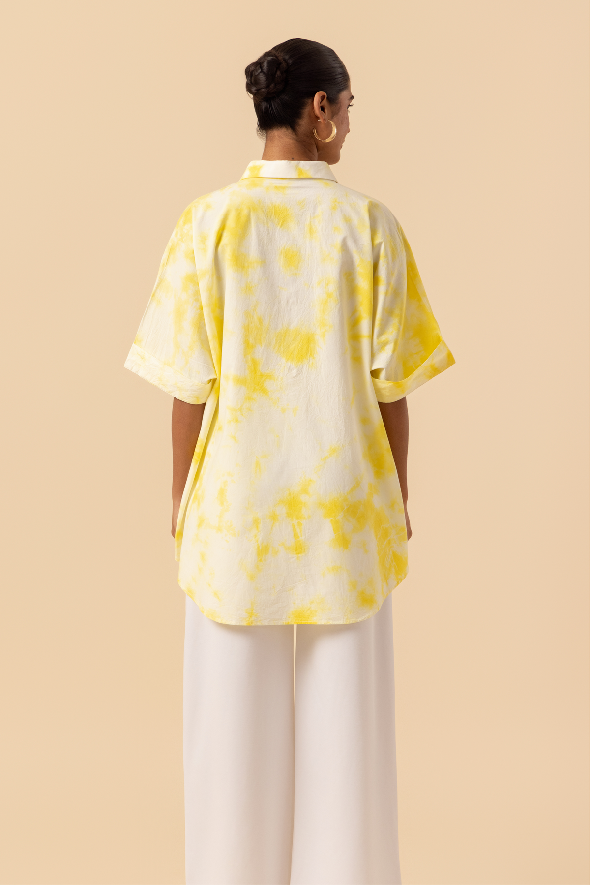 Back side view of a model poses confidently in a yellow tie-dye oversized shirt and white wide-leg pants against a soft beige background.