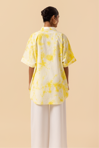 Back side view of a model poses confidently in a yellow tie-dye oversized shirt and white wide-leg pants against a soft beige background.