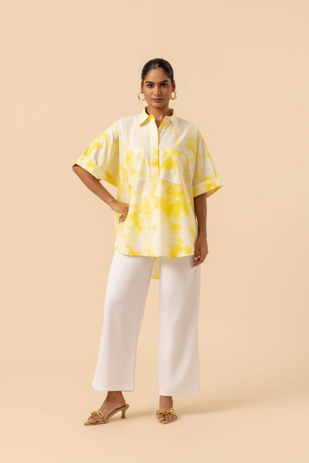 A model poses confidently in a yellow tie-dye oversized shirt and white wide-leg pants against a soft beige background.