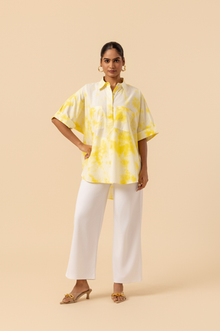 A model poses confidently in a yellow tie-dye oversized shirt and white wide-leg pants against a soft beige background.