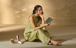 A woman sits barefoot on a beige floor, reading a book, wearing a sleeveless light olive green linen co-ord set. A pair of nude high heels rests beside her against a warm, textured beige backdrop with soft shadow overlays.