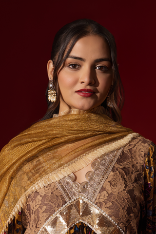 Closeup view of a woman stands confidently in a colorful, embellished regal printed velvet kurta set with a flowing golden dupatta against a vibrant red backdrop.