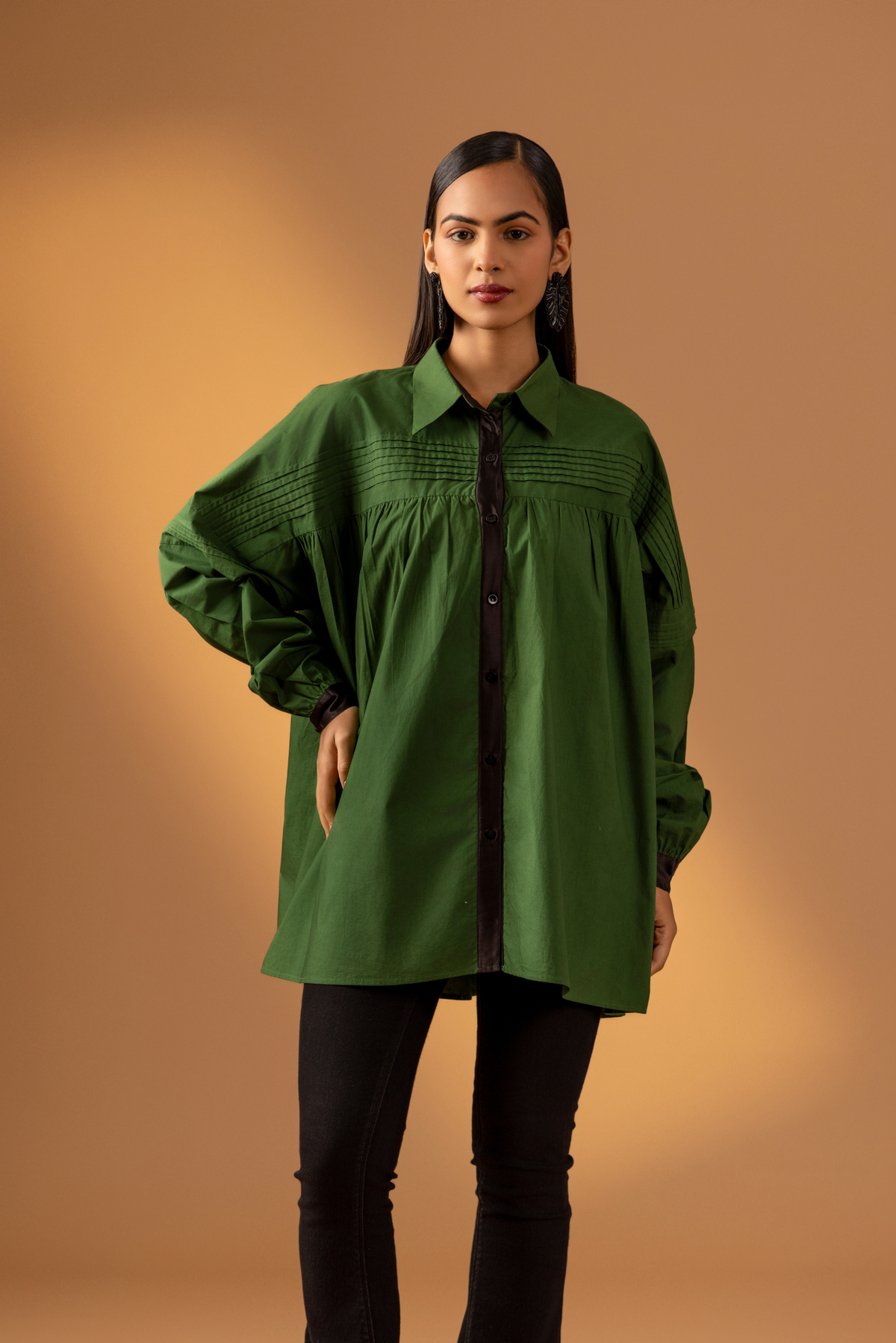 A woman stands against a warm beige background, wearing a loose green shirt with a black button front and black flared pants .