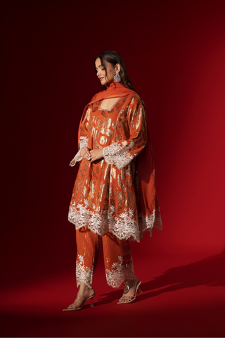 Side view of a woman poses in a orange ethnic  shimmer kurta set outfit with intricate white embroidery against a vibrant red backdrop.