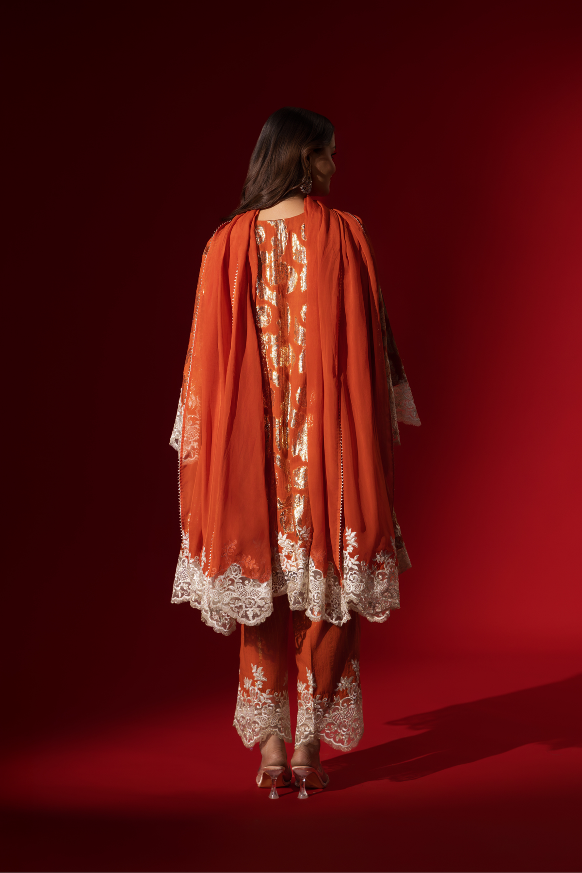 Back view of a woman poses in a orange ethnic  shimmer kurta set outfit with intricate white embroidery against a vibrant red backdrop.