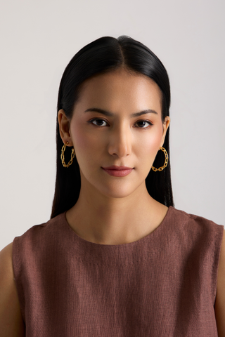 A woman with long dark hair wearing textured gold hoop earrings and a sleeveless brown top, photographed against a plain light gray background.