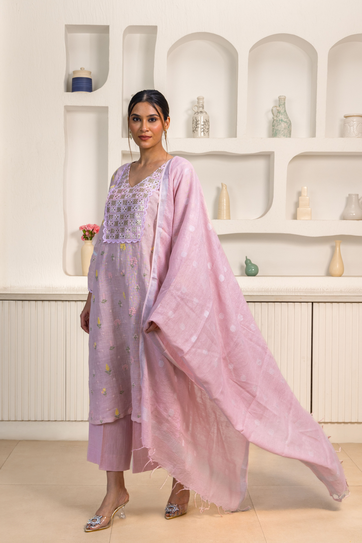 A woman wears a pastel pink traditional outfit adorned with delicate embroidery, standing against a minimalist, elegant backdrop.