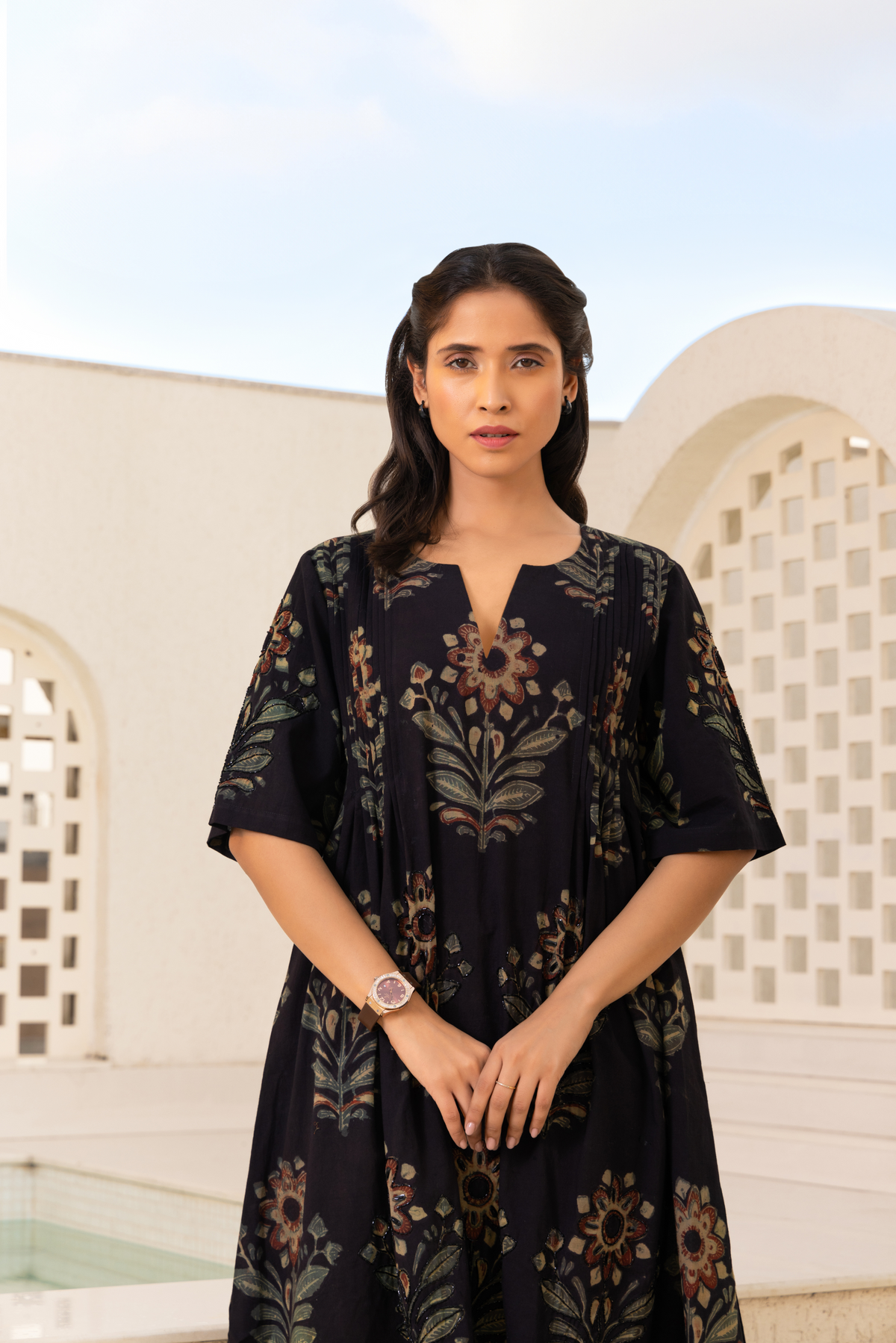 Closeup View of a woman wearing a black traditional outfit with floral embroidery and a golden hemline, paired with striped pants and black sandals, standing outdoors against a light-colored architectural backdrop and a blue sky.