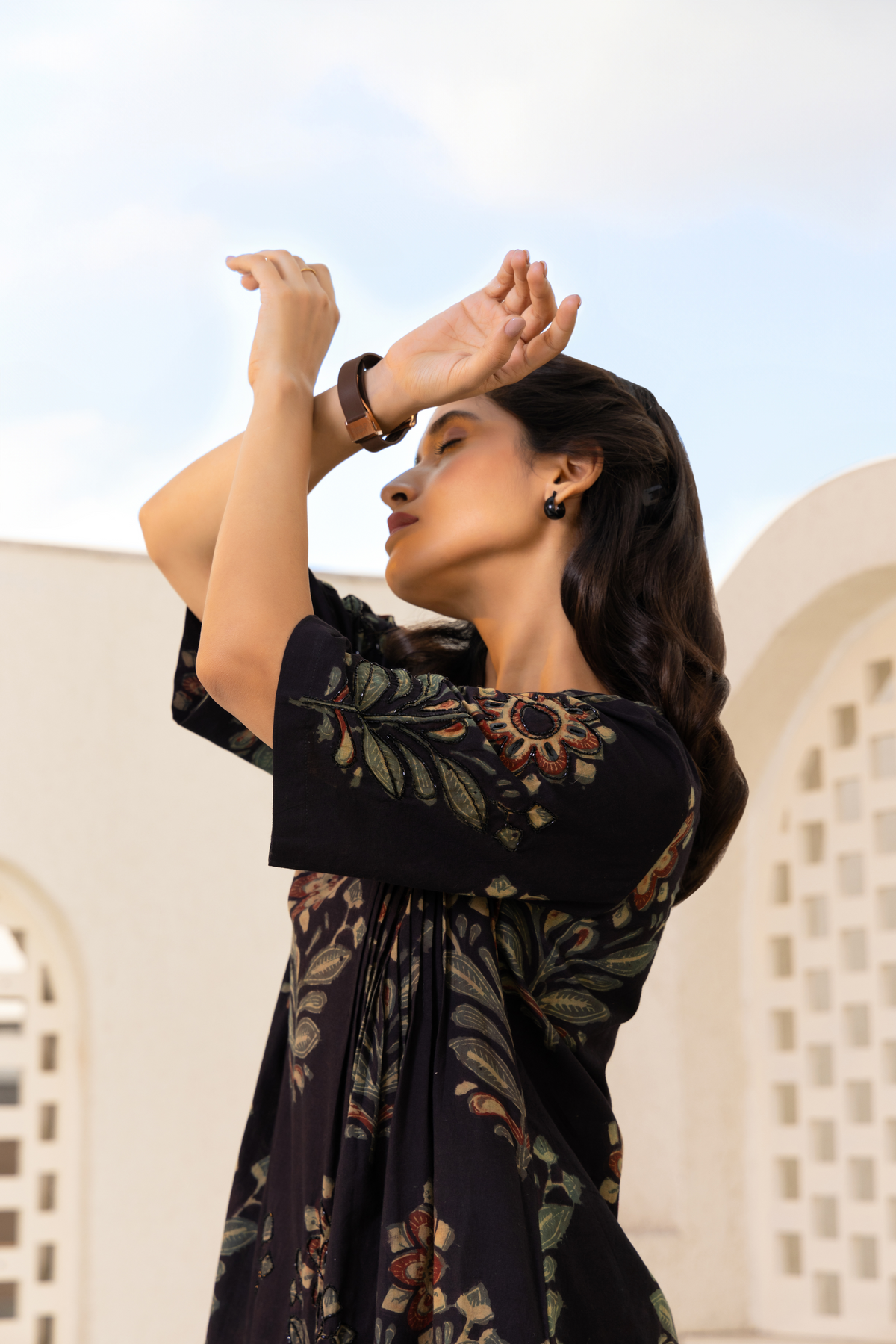 A woman wearing a black floral-printed dress poses gracefully with her arms raised against a minimalistic architectural background.