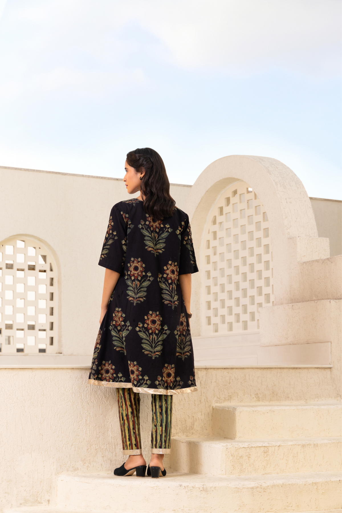 Back View of a woman wearing a black traditional outfit with floral embroidery and a golden hemline, paired with striped pants and black sandals, standing outdoors against a light-colored architectural backdrop and a blue sky.
