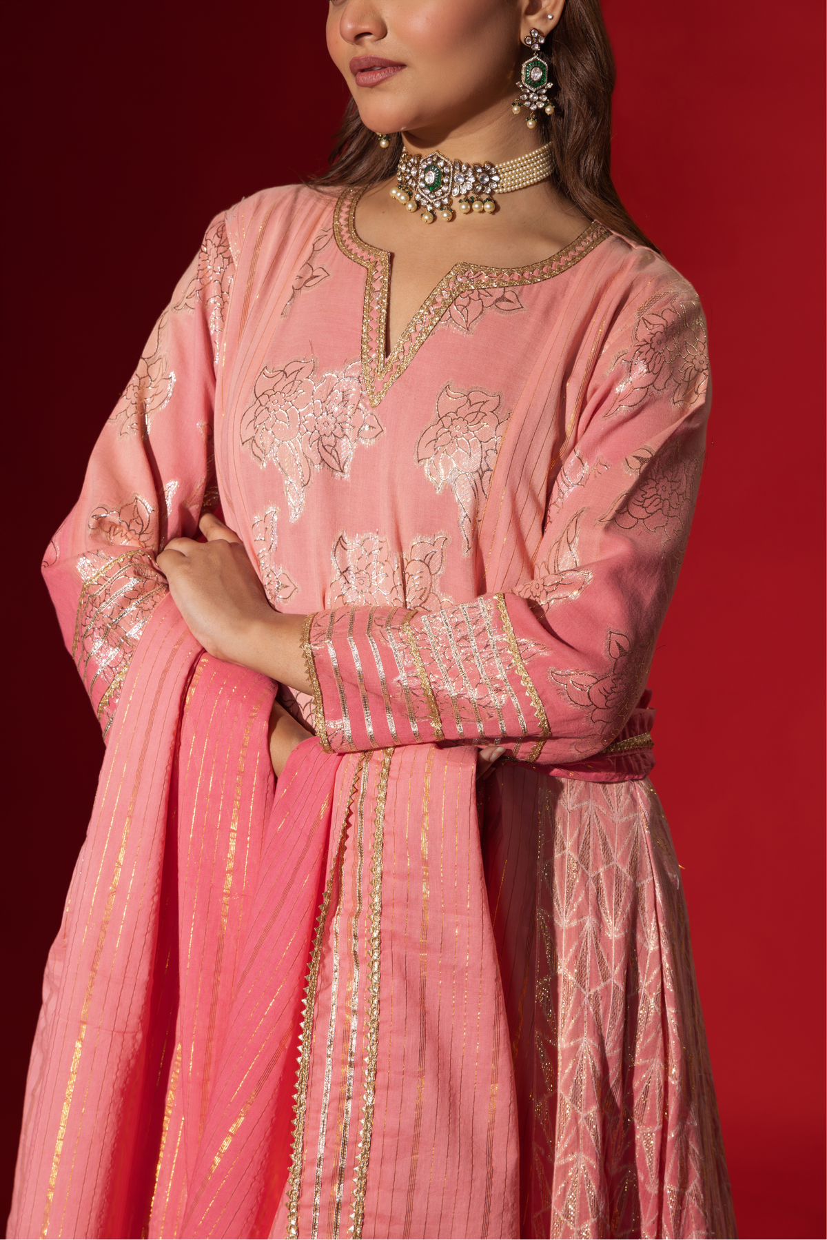 Closeup view of a model in a flowing pink ethnic outfit adorned with intricate golden patterns, standing against a deep red backdrop.
