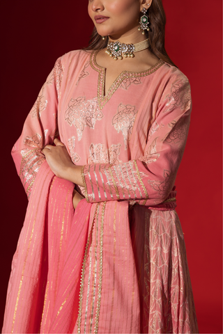 Closeup view of a model in a flowing pink ethnic outfit adorned with intricate golden patterns, standing against a deep red backdrop.