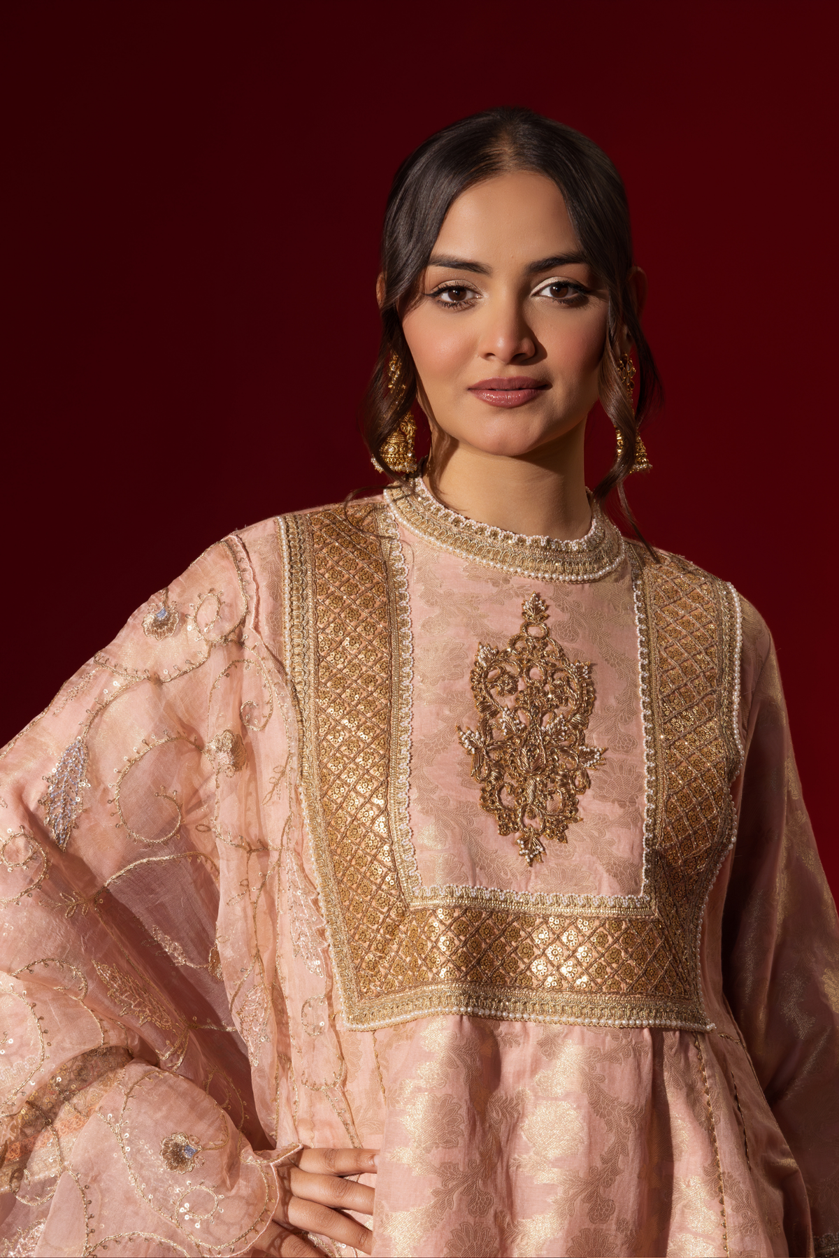 Closeup view of a woman wearing an elegant pink embroidered sequin brocade kurta set with gold accents and a sheer dupatta, set against a rich red background